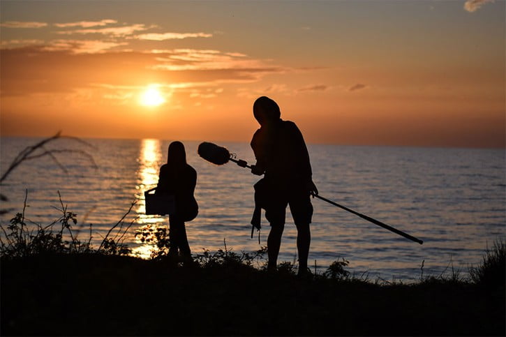 Tom Wedekind und Antonia Schmitz halten Filmklappe und Mikrofon im Sonnenuntergang am Meer.