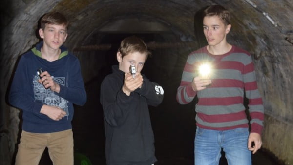 Drei Jungen stehen mit Taschenlampen in einem modrigen Tunnel. Der Mittlere zielt mit einer Pistole.