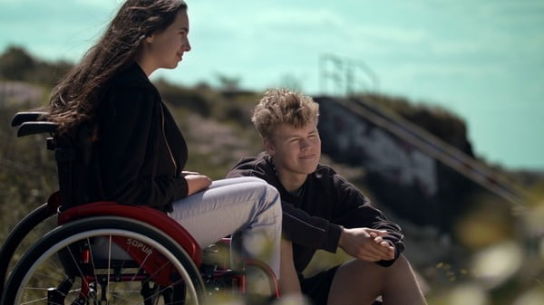 Ein Mädchen im Rollstuhl und ein Junge sitzen nebeneinander in der Natur und sehen in die Ferne.