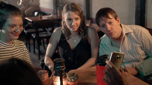 Drei Menschen in einer Bar sehen interessiert auf ein Handy, das ihnen entgegengehalten wird.
