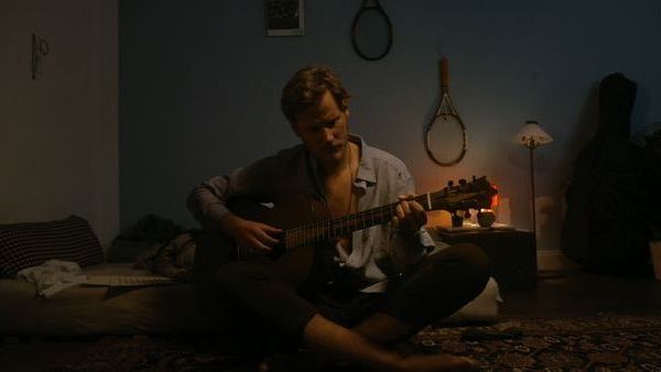 Ein Mann spielt Gitarre, während er auf einer Matratze in einem dunklen Raum sitzt.